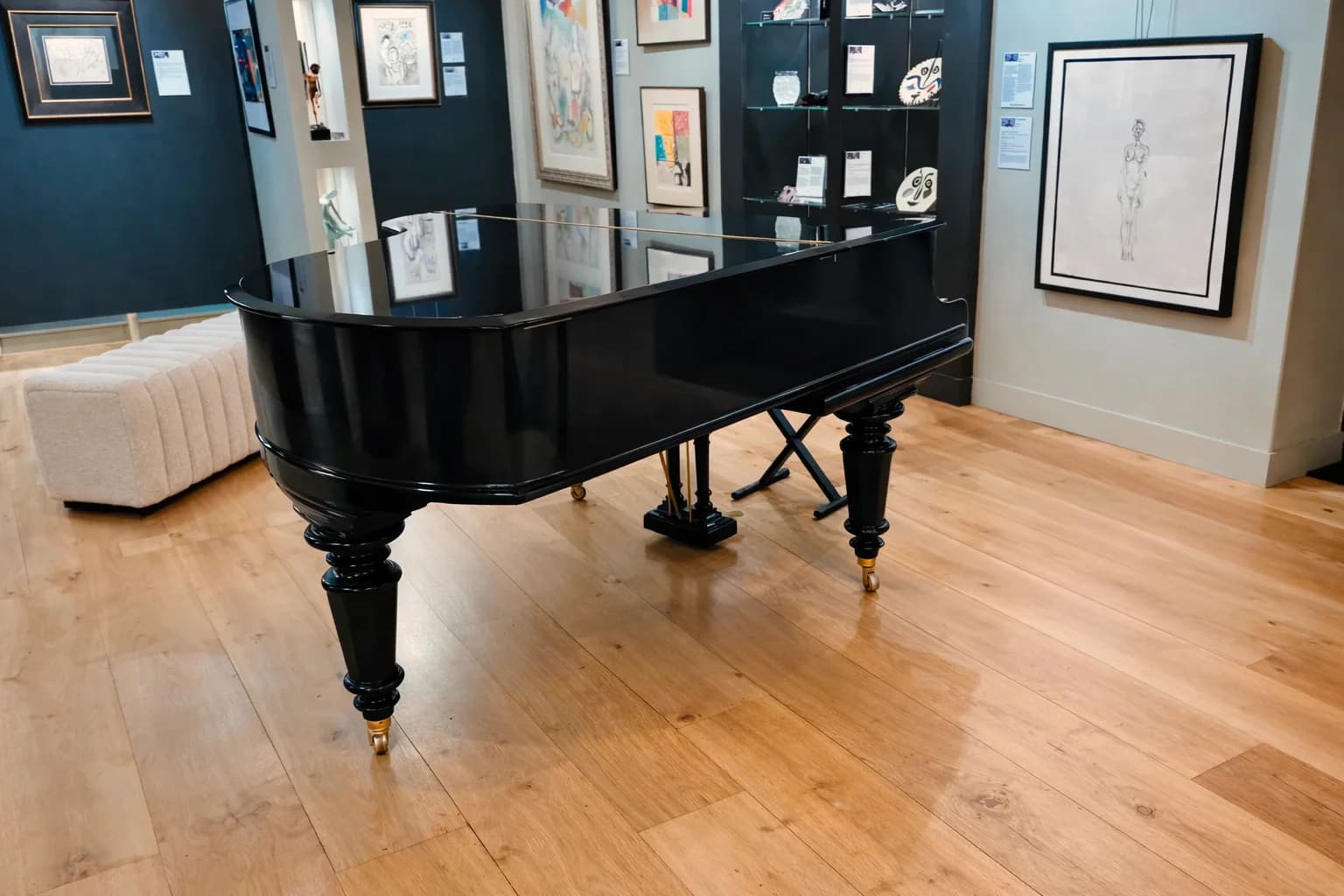 Polished black grand piano on light wood floors in an art gallery with framed artwork.
